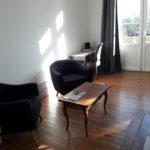 Cosy living room with two black armchairs, an old wooden coffee table, a beautiful desk, wooden floors and a large French window opening onto the terrace